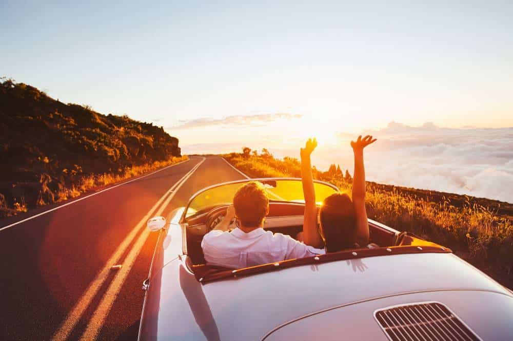 Un jeune couple dans une voiture décapotable sur une route de campagne avec un couché de soleil.